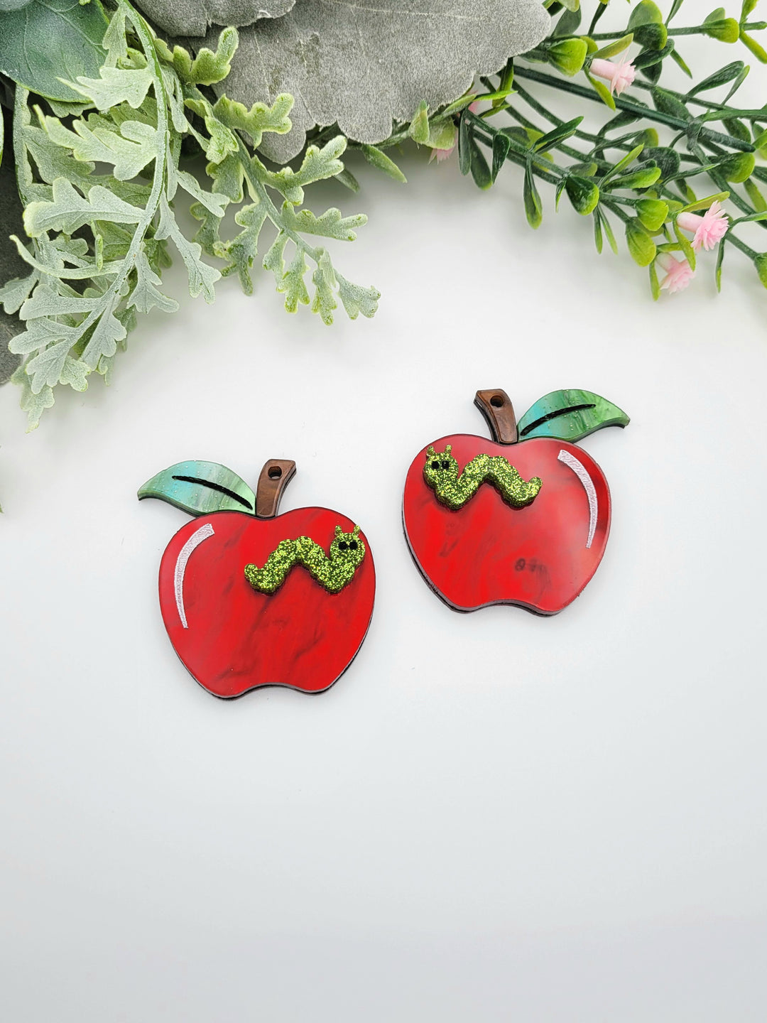 two red apple-shaped objects with green leaves and a small green caterpillar on them, placed on a white background.